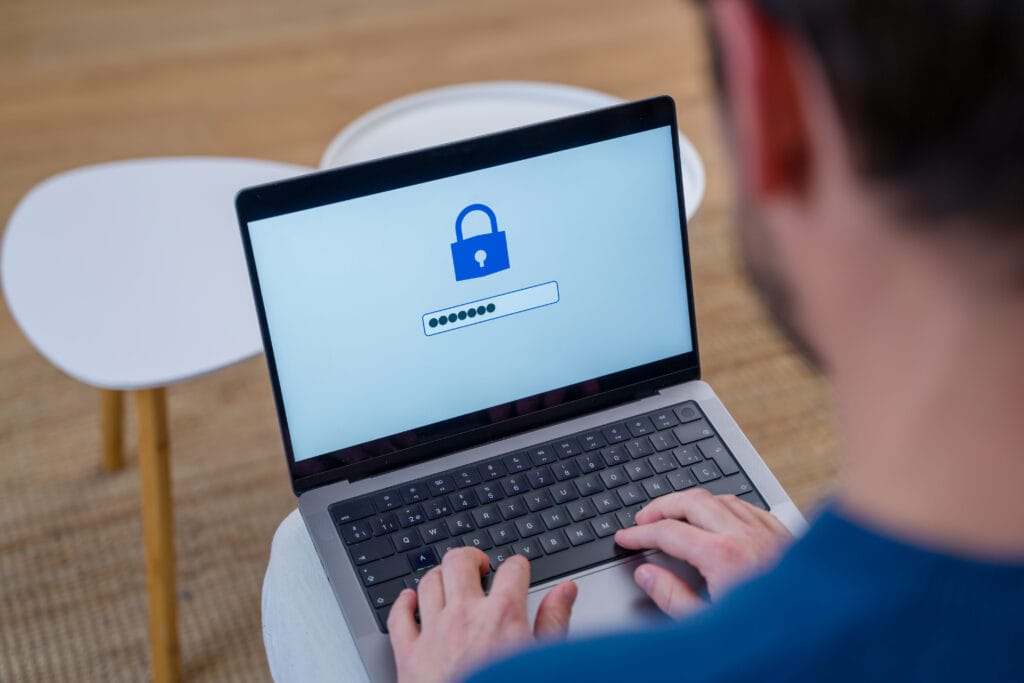 Looking over a man's shoulder toward his laptop with a lock showing on screen while he enters a password.